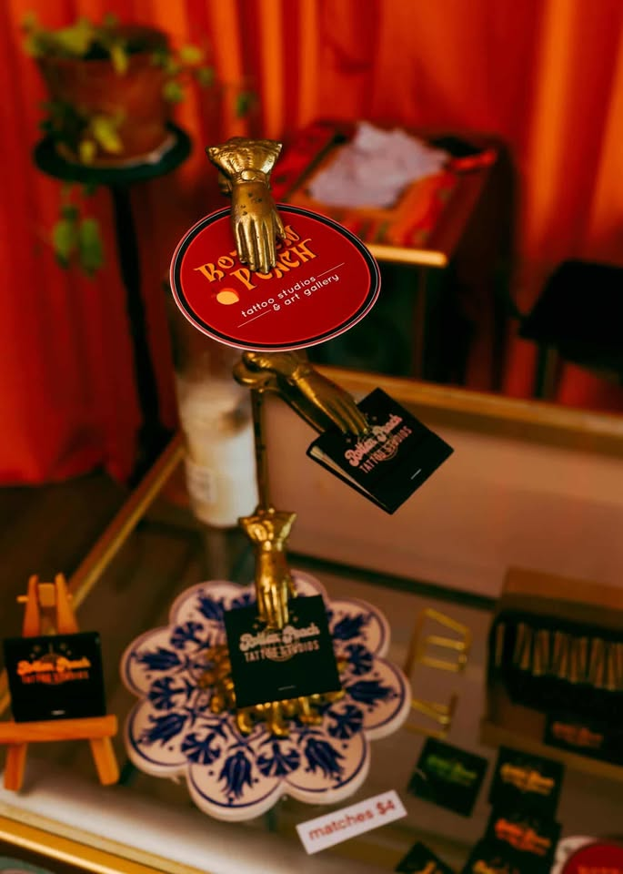 Rotten Peach Studios red-oval logo on a gold hand stand with stickers and matchbooks